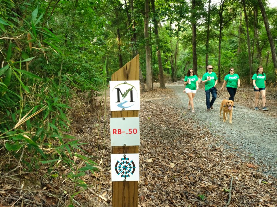 Four people in green shirts walk towards the viewer with a golden retriever, along a gravel trail. Signpost with trail information in foreground, lush forest in background.